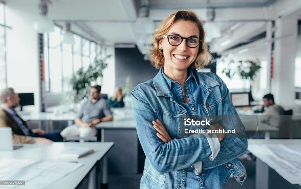 Happy female designer standing in office Successful creative female standing in office with colleagues in background. Happy woman executive standing in office with arms crossed. Women Stock Photo