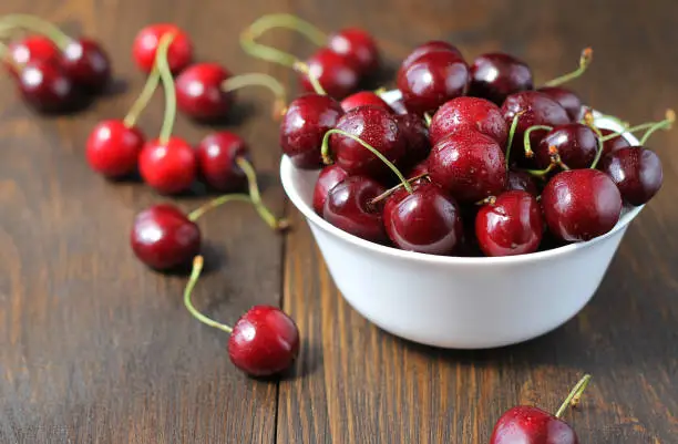 Sweet cherries in the white bowl on the wooden table. Scattered cherries on the table.