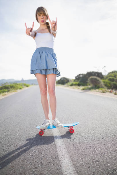 Trendy woman making rock and roll gesture while balancing on her skateboard on a deserted road