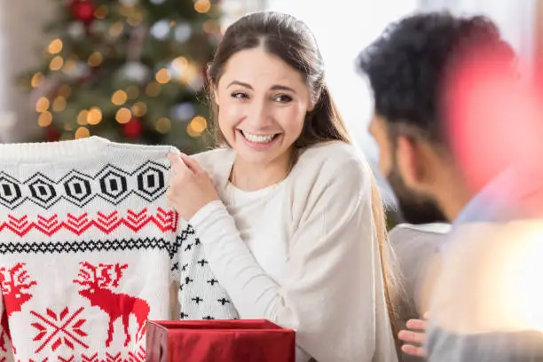 A young woman both smiles and grimaces as she holds up a Christmas sweater she has just opened for her unrecognizable friend to see.  She is sitting on the couch in her living room.