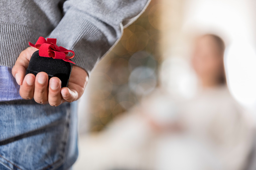 This is a closeup with copy space of an unrecognizable young man with focus on a ring box as he holds it behind his hip, getting ready to propose to his unidentifiable girlfriend in the background.