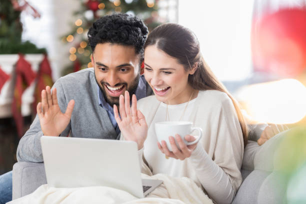 Young couple video chats with family at Christmas A smiling young couple wave at their laptop while she sits on their couch and he crouches beside her in their living room at Christmas. She is holding a coffee cup as they video conference with out of town family. asian women in stockings stock pictures, royalty-free photos & images