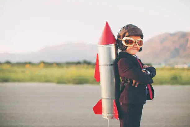 A young business minded boy is wearing a business suit, space helmet with a rocket strapped to his back. His hands are on his hips and he is smiling at the camera standing on blacktop. Plenty of copy space for your innovative type. Image taken in Utah, USA.