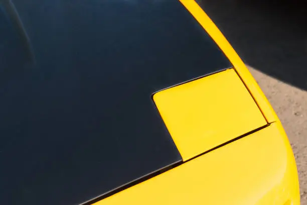 Sports car hood of a yellow and black color against the dark shadow and bright brown spot on the ground. Play of light and shadows, colors and shapes. Automotive abstract.
