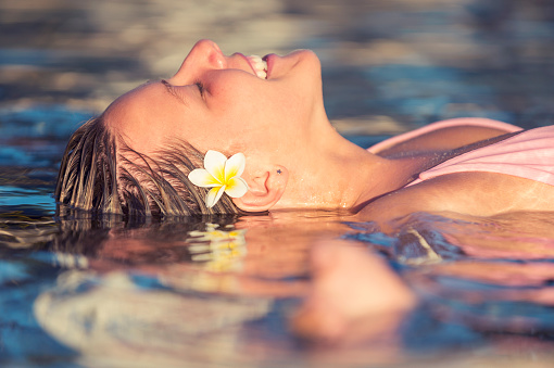 Portrait of Woman relaxing floating in the water. Light is sunset . She has a frangipani flower in her hair. She could be in a health spa or tourist resort. She is smiling and happy.