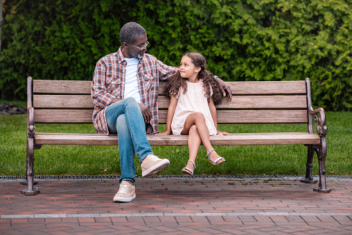 adorable african american girl and her grandfather sitting on bench in park
