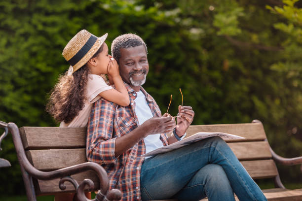 nieto susurrándole a su abuelo sonriente sentado sobre un banco en el parque - whispering grandparent child grandfather fotografías e imágenes de stock