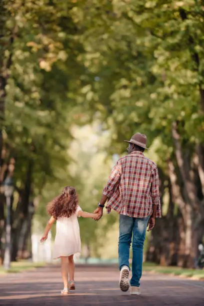 Photo of girl holding hands with grandfather and walking in green alley