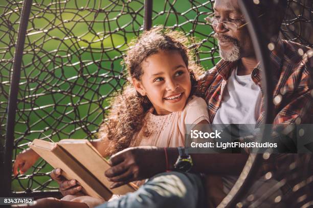 Granddaughter Reading Book While Sitting In Swinging Hanging Chair With Grandfather Stock Photo - Download Image Now