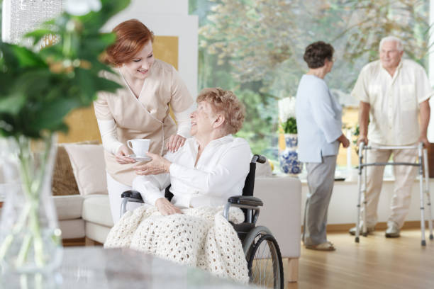 Friendly nurse gives a cup of tea Friendly nurse gives a cup of tea to disabled woman on wheelchair in senior home common room stock pictures, royalty-free photos & images