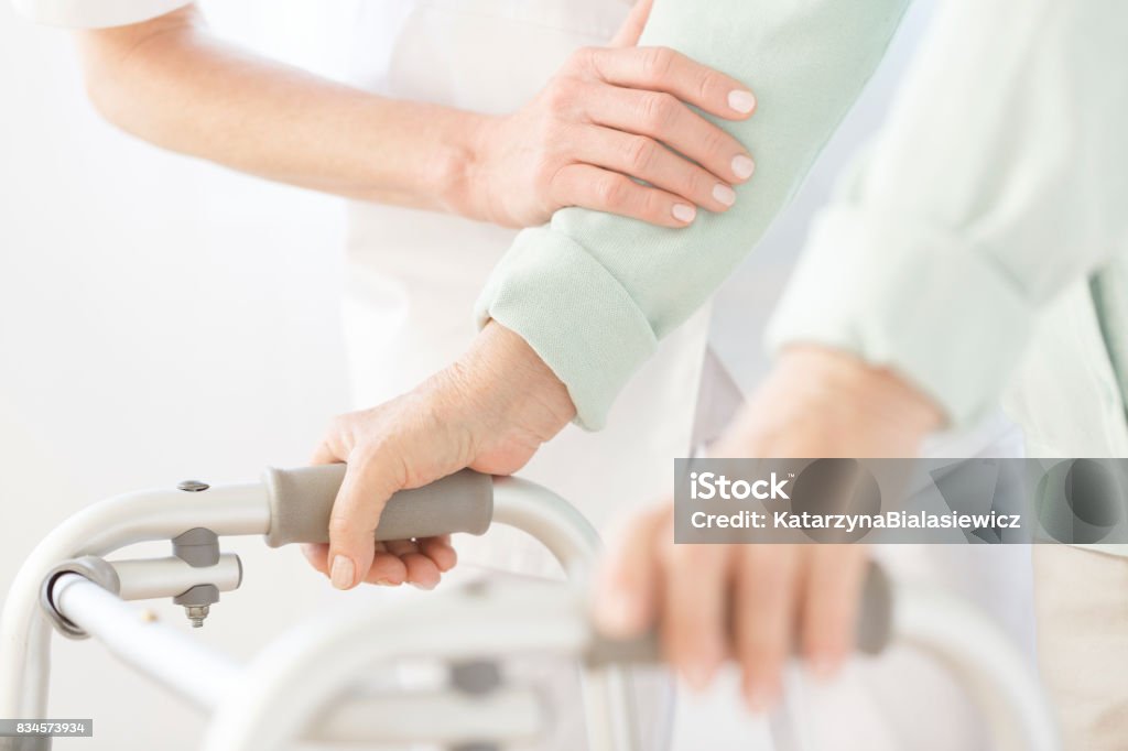 Assistant in white uniform Assistant in white uniform hold patient's arm while he uses walking frame Home Caregiver Stock Photo