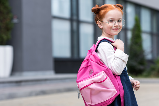 cute little redhead schoolgirl in eyeglasses holding backpack and smiling at camera