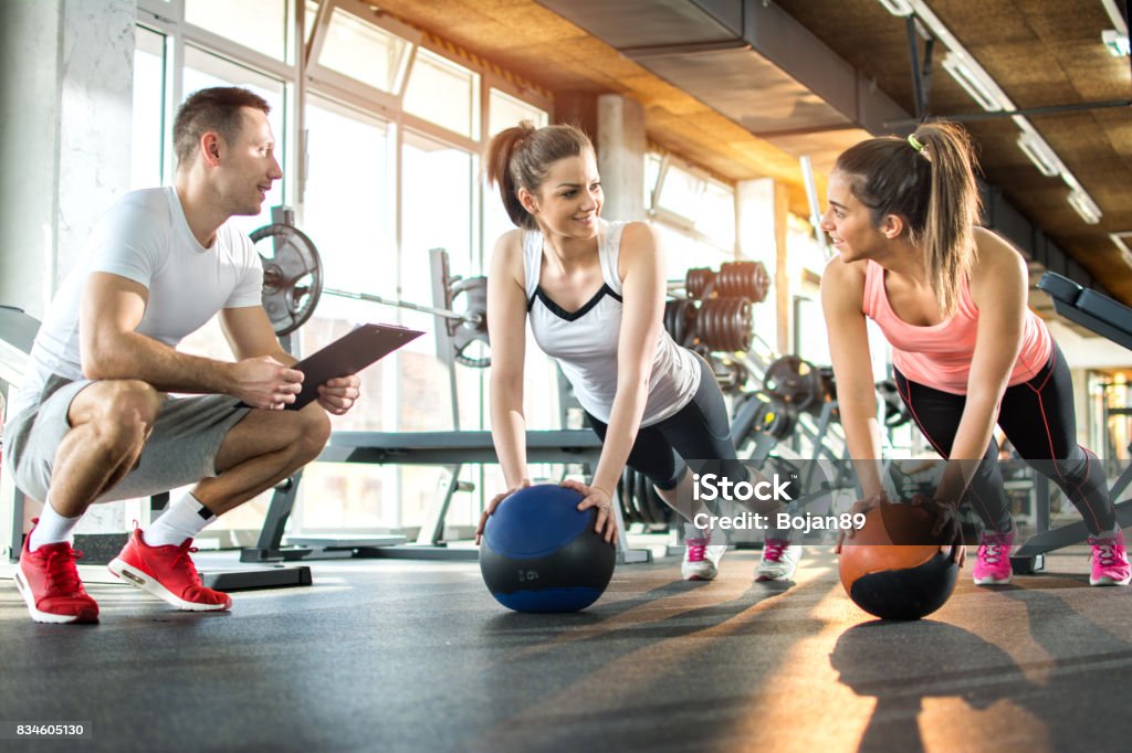 Two sporty girls exercising with fitness balls while their fitness instructor tracking the progress on clipboard. Fitness Instructor Stock Photo