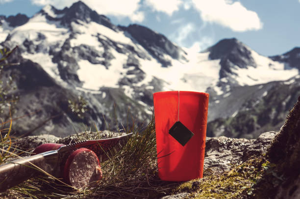 Orange plastic mug with strong tea and tea bag on a snowy winter background. Tourist knife cuts smoked sausage.
