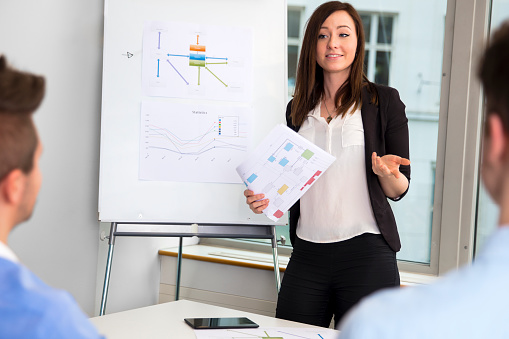 Confident young businesswoman holding chart while communicating with colleagues in office