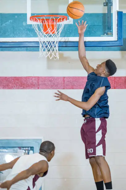 A group of young men are playing basketball indoors in a gym. They are wearing athletic clothes and shoes. One man is leaping up and trying to sink the ball.