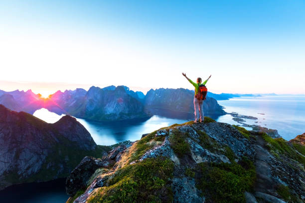Hiker enjoying midnight sun during arctic summer, Reine, Lofoten, Norway Happy woman hiker enjoying scenic view of midnight sun at the top of Reinebringen hike above Reine village in the Lofoten archipelago during arctic summer, Norway lofoten and vesteral islands photos stock pictures, royalty-free photos & images