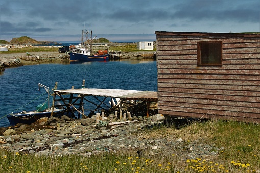 Fishing shed and stage, Ferryland, Newfoundland and Labrador, Canada. A small boat is tied up to the pier. a larger fishing bat sits at anchor across the bay.