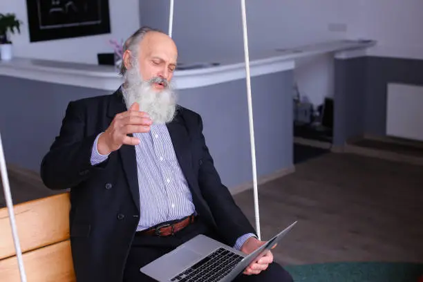 Old man holds laptop in hands and emotionally tells employees story or discusses at expense of questions in work, learns to understand programs and sits on suspended wooden bench on cables in comfortable recreation area of modern company. Elderly man with long gray beard of European appearance dressed in black classic suit and white shirt in blue stripes. Concept of elderly businessman in office, combining work during pension, training and assistance in mastering modern technology, lunch break after work.