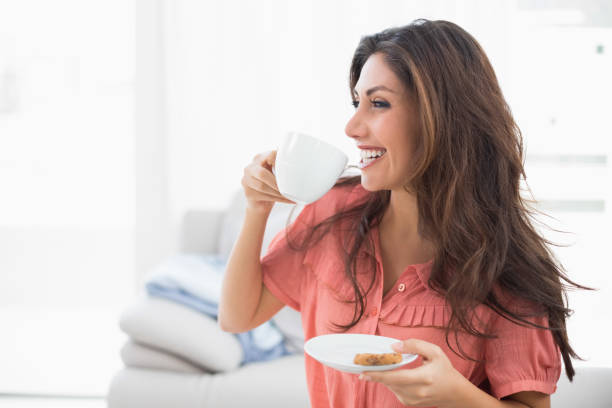 istock Happy brunette sitting on her sofa holding cup and saucer with a cookie 835149608