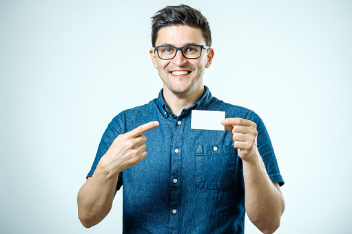 Casual young businessman holding and pointing business card. Isolated on gray