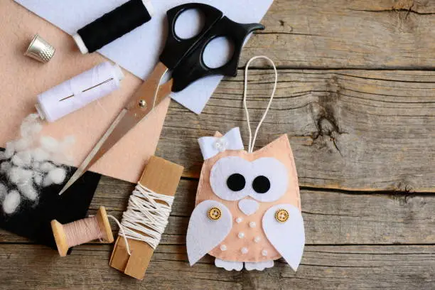 Photo of Lovely owl Christmas tree ornament made from felt. Scissors, thread, needle, thimble, felt sheets, waxed cord on an old wooden table. Creating felt owl crafts. Children sewing craft concept. Top view