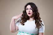 Surprised plus size fashion model, fat emotional woman on beige background, overweight female body