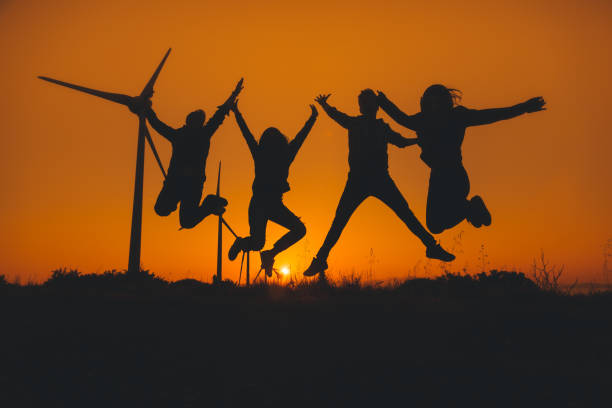 Group of happy four young people jumping on wind farm area outdoor at sunset time