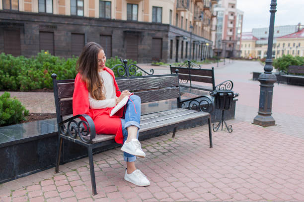 une jolie fille avec longs cheveux bruns et un sourire de la dent blanche est assis sur un banc et écrit ses pensées sur le fond urbain dans un carnet rouge. elle est vêtue d’un chandail blanc et bleu jeans - paper document pen long hair photos et images de collection