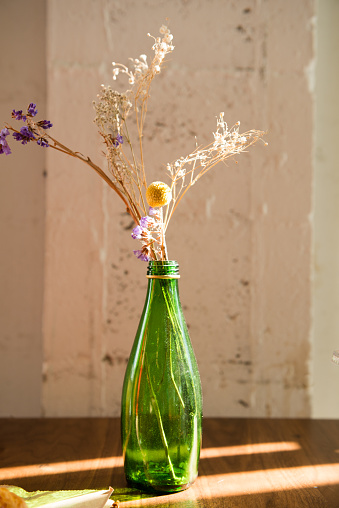 This is a vertical, color photograph of a repurposed, glass bottle being upcycled as a vase for a table display. Natural sunlight shines across the table.