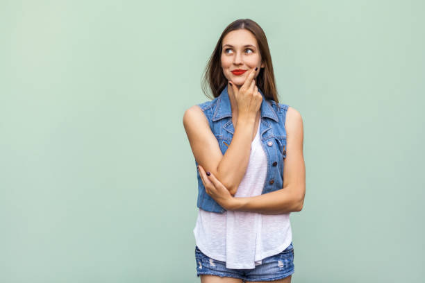 joyeux joyeuse adolescente avec taches de rousseur, style décontracté blanc veste t shirt et jeans, levant, de penser et de toucher son visage et bénéficiant d’un, bon jour et gratuit le temps à l’intérieur. studio, coup, fond vert clair - face time photos et images de collection