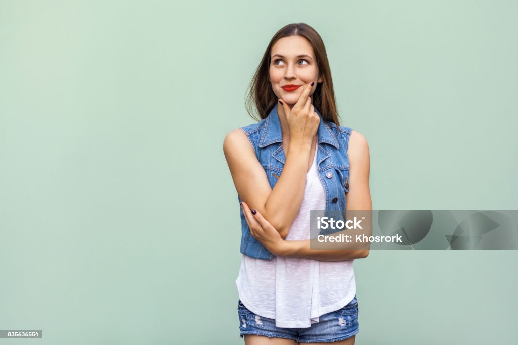 Happy cheerful teenage girl with freckles, casual style white t shirt and jeans jacket, looking up, thinking and touching her face and a, enjoying good day and free time indoors. Studio shot, light green background Women Stock Photo