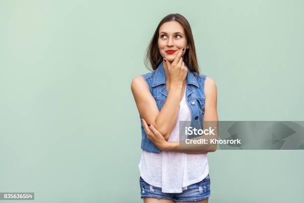 Happy Cheerful Teenage Girl With Freckles Casual Style White T Shirt And Jeans Jacket Looking Up Thinking And Touching Her Face And A Enjoying Good Day And Free Time Indoors Studio Shot Light Green Background Stock Photo - Download Image Now