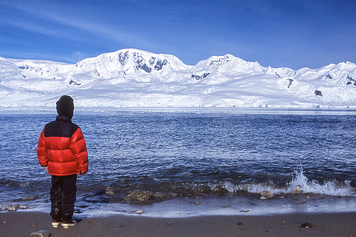 Young boy on antarctica beach looking out over harbor at distant snow covered mountain peaks.








