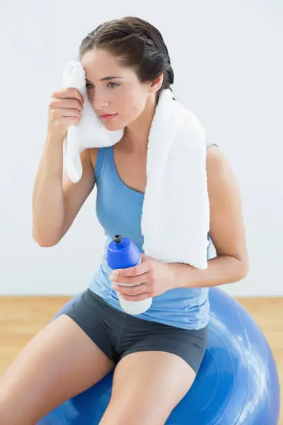 Tired young woman with towel around neck and waterbottle sitting on exercise ball