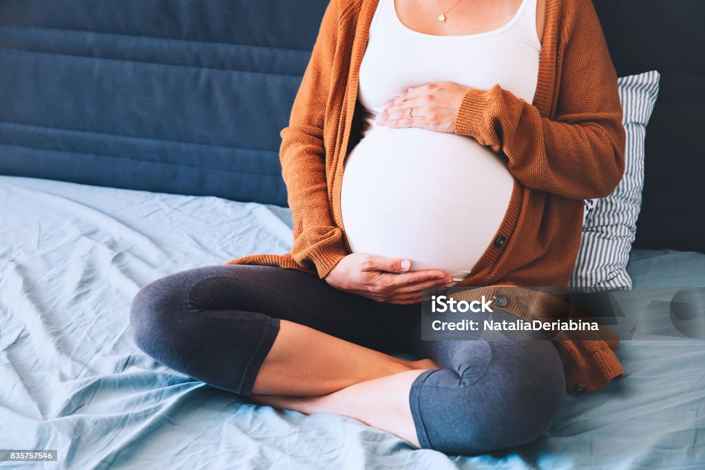 Schöne schwangere Frau sitzen am Bett und hält die Hände auf den Bauch im Schlafzimmer zu Hause. - Lizenzfrei Abwarten Stock-Foto