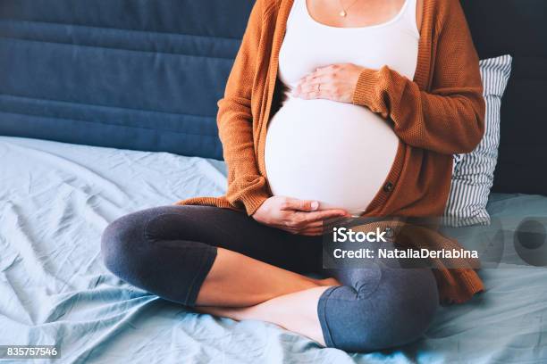 Beautiful Pregnant Woman Sitting At Bed And Holds Hands On Belly In Bedroom At Home Stock Photo - Download Image Now