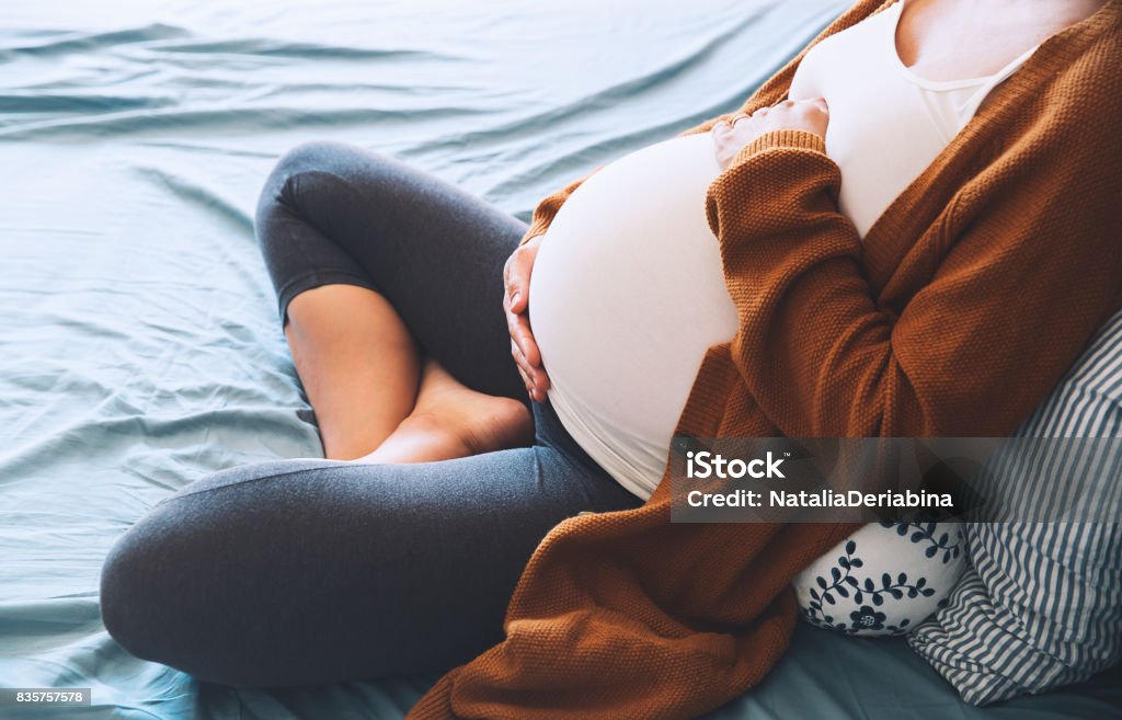 Beautiful pregnant woman sitting at bed and holds hands on belly in bedroom at home. Beautiful pregnant woman sitting at bed and holds hands on belly in bedroom at home. Young mother waiting of a baby. Pregnancy, maternity and expectation image. Close-up, indoors. Pregnant Stock Photo