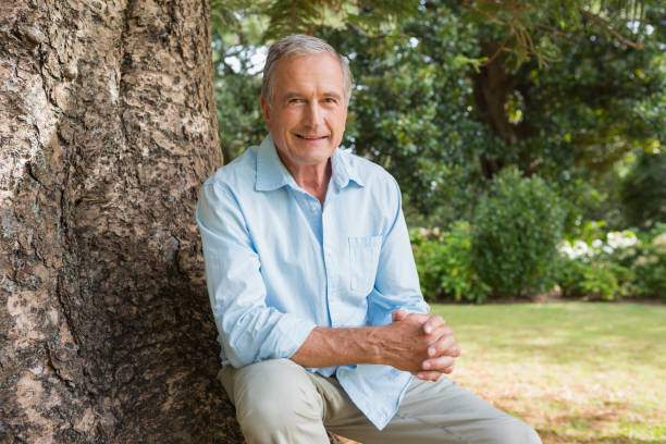 Cheerful mature man sitting on tree trunk Cheerful mature man sitting on tree trunk smiling at camera in the park 65 69 years stock pictures, royalty-free photos & images