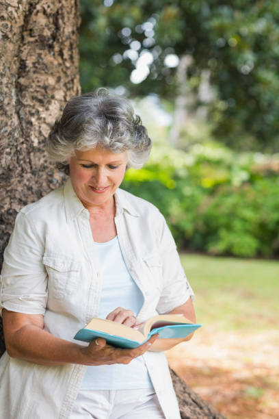 istock Cheerful mature woman reading book leaning on tree trunk 835772358