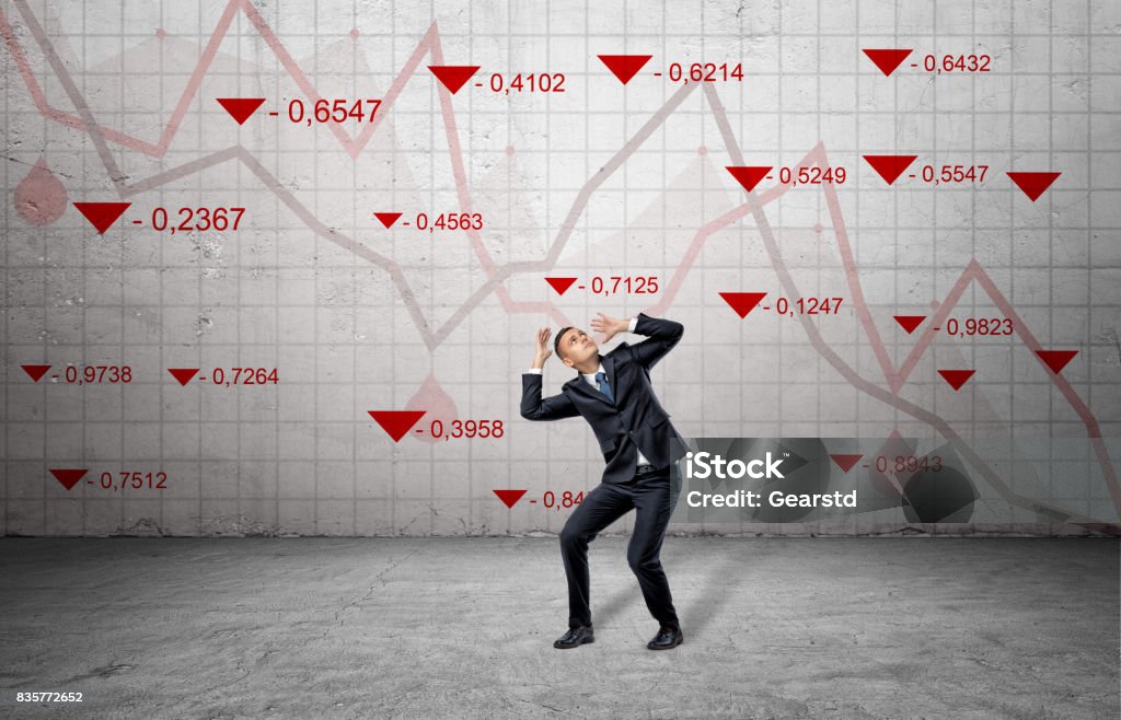 A scared businessman cowers near a concrete wall with red stock market indexes and falling statistic lines A scared businessman cowers near a concrete wall with red stock market indexes and falling statistic lines. Financial markets. Bear trading. Losing money. Stock Market and Exchange Stock Photo