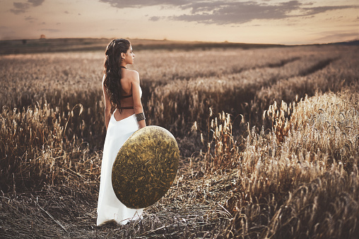 Back view of brunette girl with long hair wearing in white dress like greece style,  holding iron shield in hand. Wife waiting husband after war, among grass in field turned and looking away.