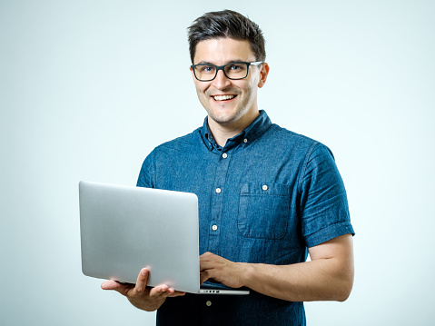 Confident young handsome man in shirt holding laptop while standing against white background