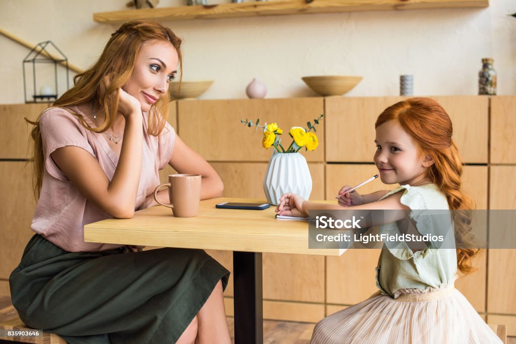 side view of smiling young mother looking at cute little daughter drawing with pencil in cafe Adult Stock Photo