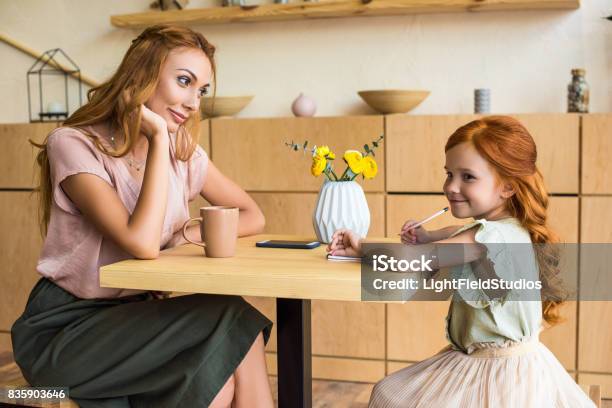 Side View Of Smiling Young Mother Looking At Cute Little Daughter Drawing With Pencil In Cafe Stock Photo - Download Image Now