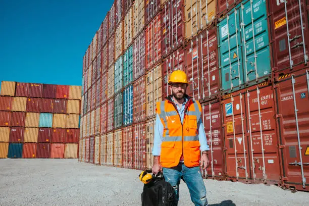 Maintanence port worker man with yellow crash helmet and worker west posing and smiling in front of colorful cargo container stacks in shipping port