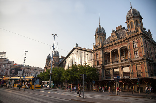 Picture of the main buidling of Nyugati train station in Budapest, with a tram in front and pedestrians crossing by