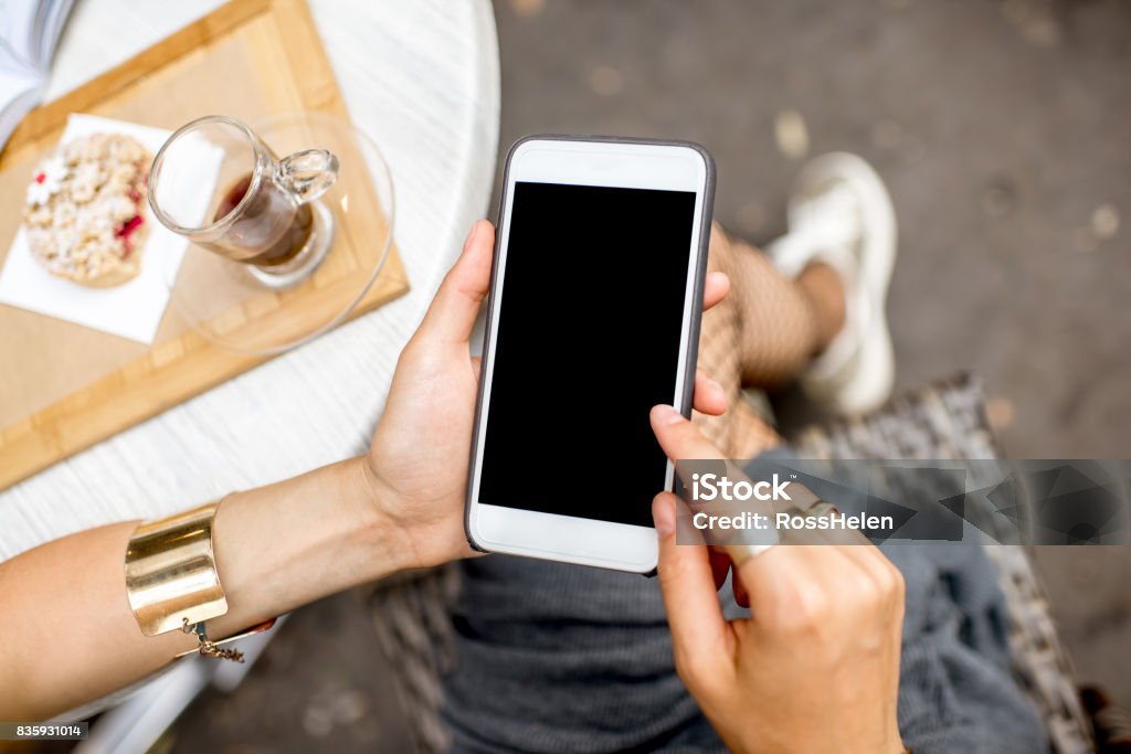 Using a smartphone at the cafe Woman holding a smart phone with empty screen sitting outdoors at the cafe with cake and coffee on the table Telephone Stock Photo