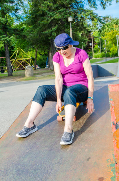 istock Senior woman wearing baseball cap on the side and sitting on skateboard while going down ramp during summer day in skate park 835934610