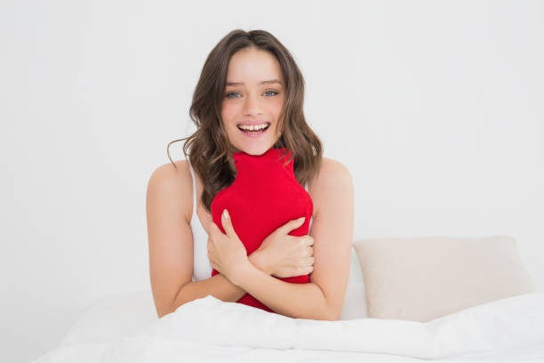Smiling woman with a hot water bottle in bed Portrait of a smiling young woman sitting with a hot water bottle in bed red cushion covers stock pictures, royalty-free photos & images